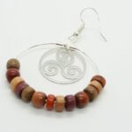 Boucles d’oreilles créoles en bois multicolore et filigrane argenté en forme de triskel, posées sur fond neutre.